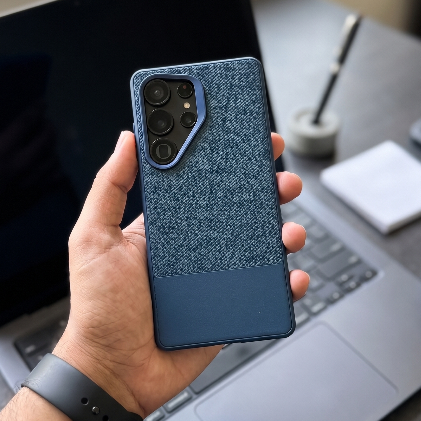 Hand holding a blue phone case in front of a laptop on a desk.