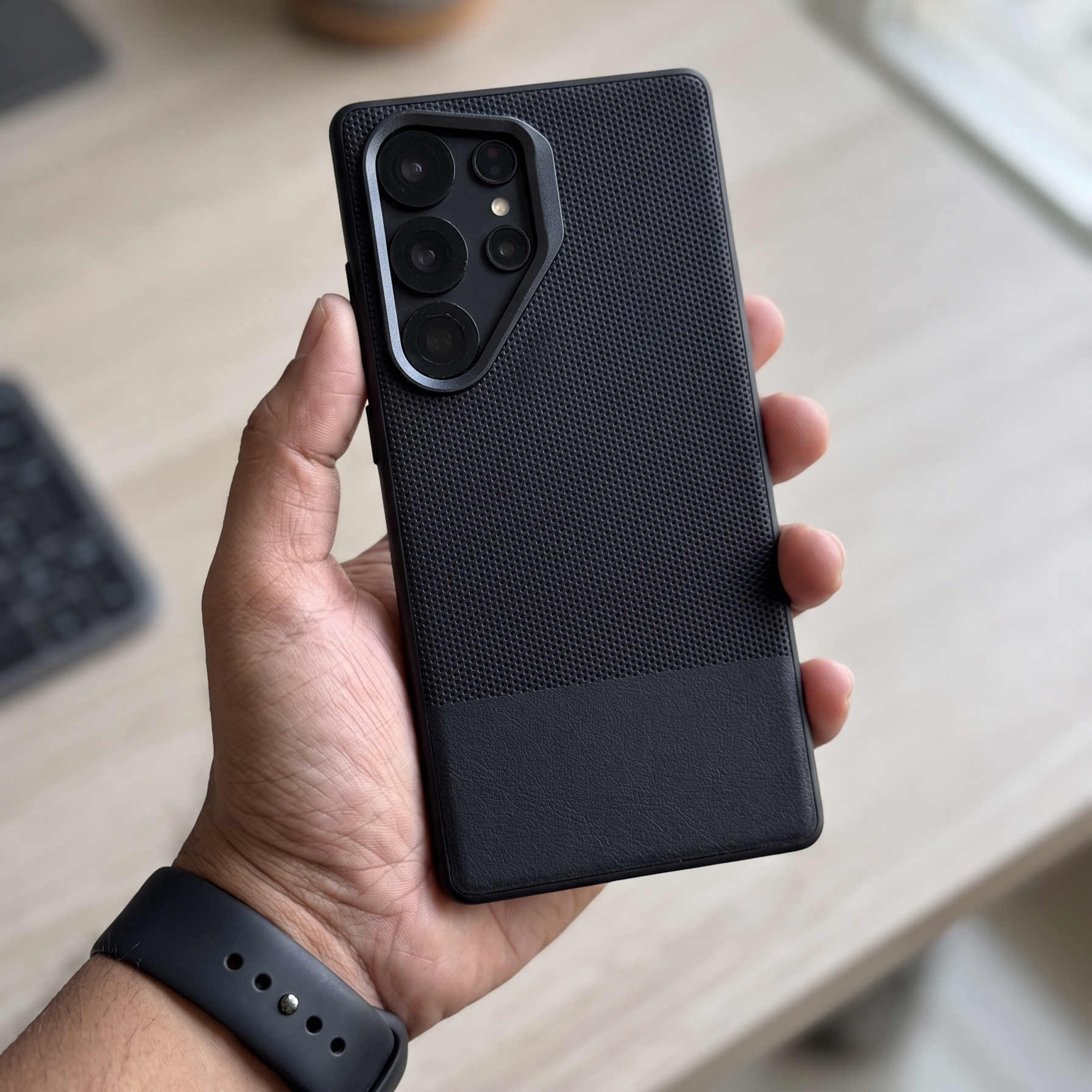 Person holding a black phone case on a desk with a cup and keyboard in the background